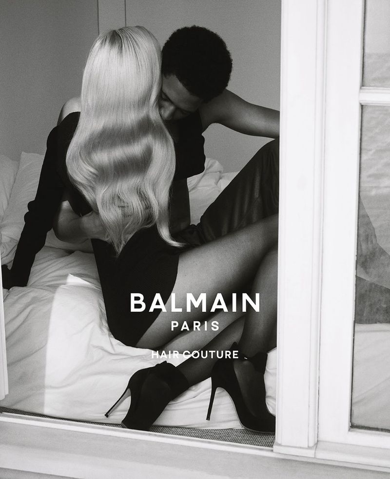 Black and white editorial image of a blonde woman and a man embracing on a bed for Balmain Hair