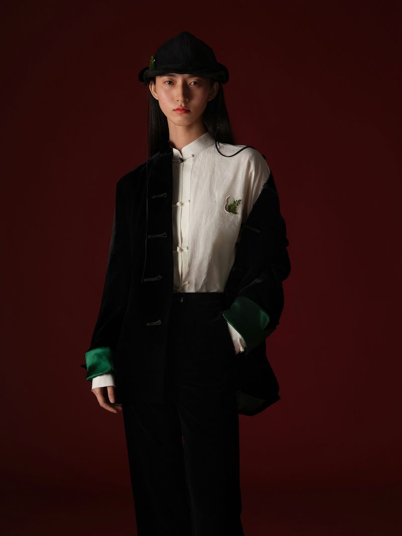 A stylish Asian woman in a black hat and suit stands against a red background, presenting an elegant fashion portrait.
