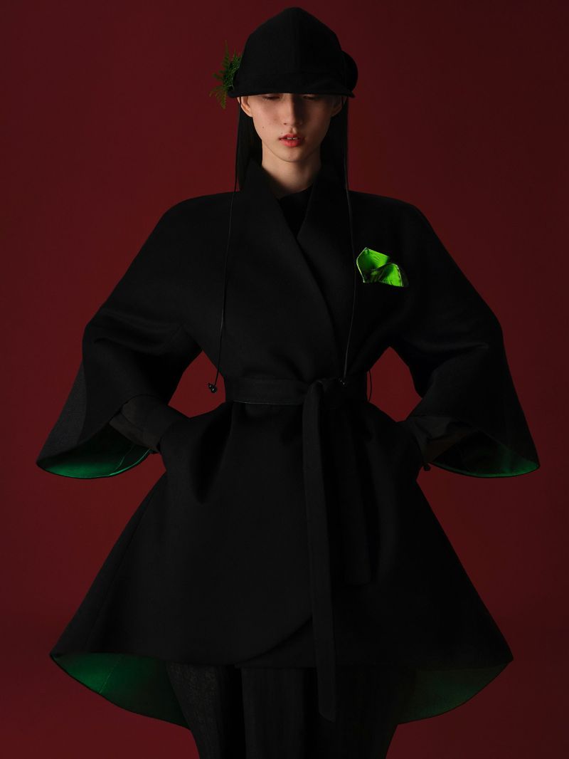 Fashion portrait of a woman wearing a black wrap coat with a green lining and a matching hat against a dark red studio backdrop.