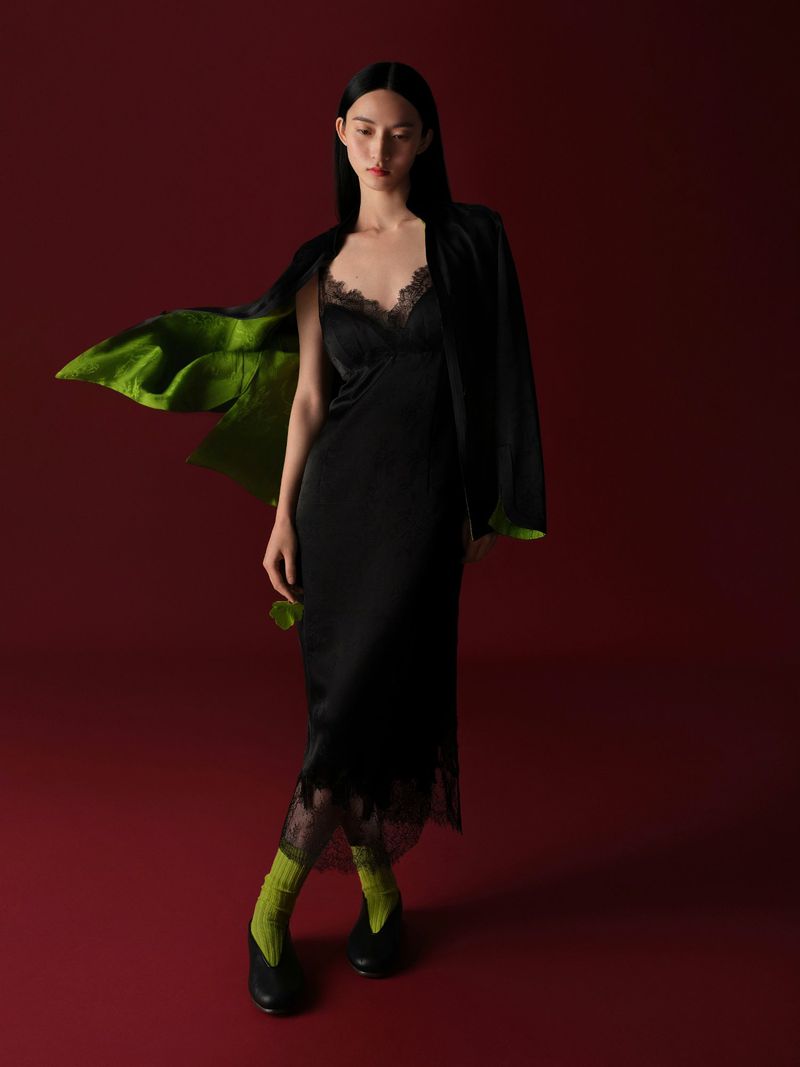 A slender adult female model poses in a long black slip dress with lace, a black jacket, and green socks against a dark red background.