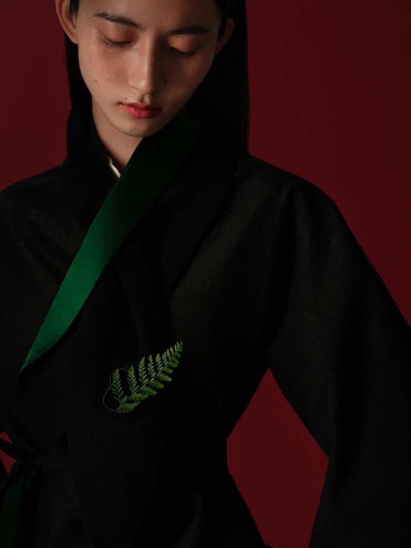 Young adult in a black kimono-style garment with a green fern embellishment, captured in a close-up studio shot against a red backdrop.