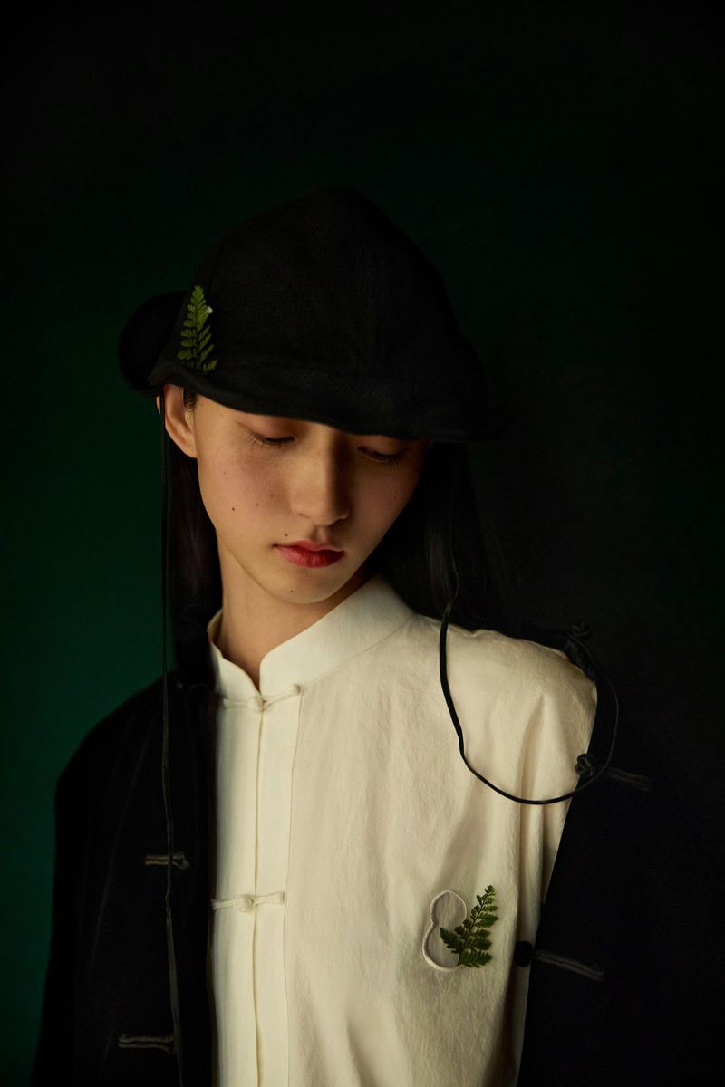 Portrait of an Asian woman in black hat and split-color top against a dark background, accented with delicate fern details.