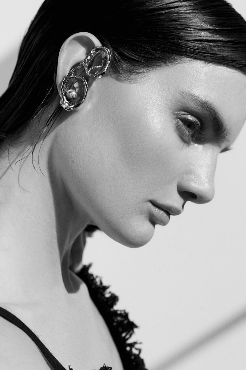 Monochrome portrait of a woman with slicked-back hair and an abstract sculptural earring, standing in front of a white background.