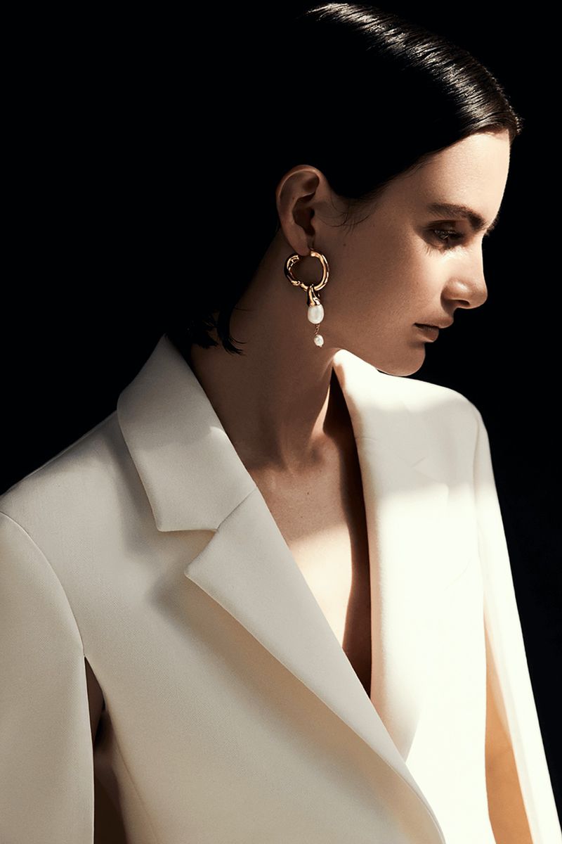 Elegant woman with dark hair and pearl drop earring wears a white blazer in a dramatic fashion portrait against a dark background.