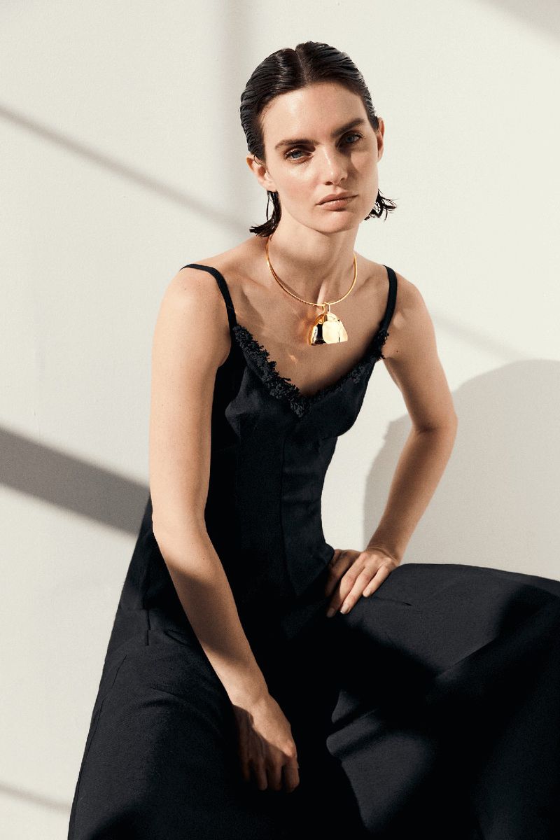 Elegant woman in a black dress and gold necklace poses against a light wall, showcasing fashion and portrait photography.