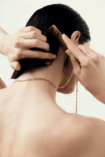 Woman with short black hair is shown from the back, combing her hair with a brown comb. She wears a gold necklace and earring.