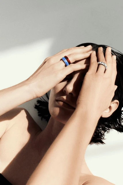 Woman poses indoors with hands on her head, wearing colourful rings in a beauty and fashion shot with a white background.
