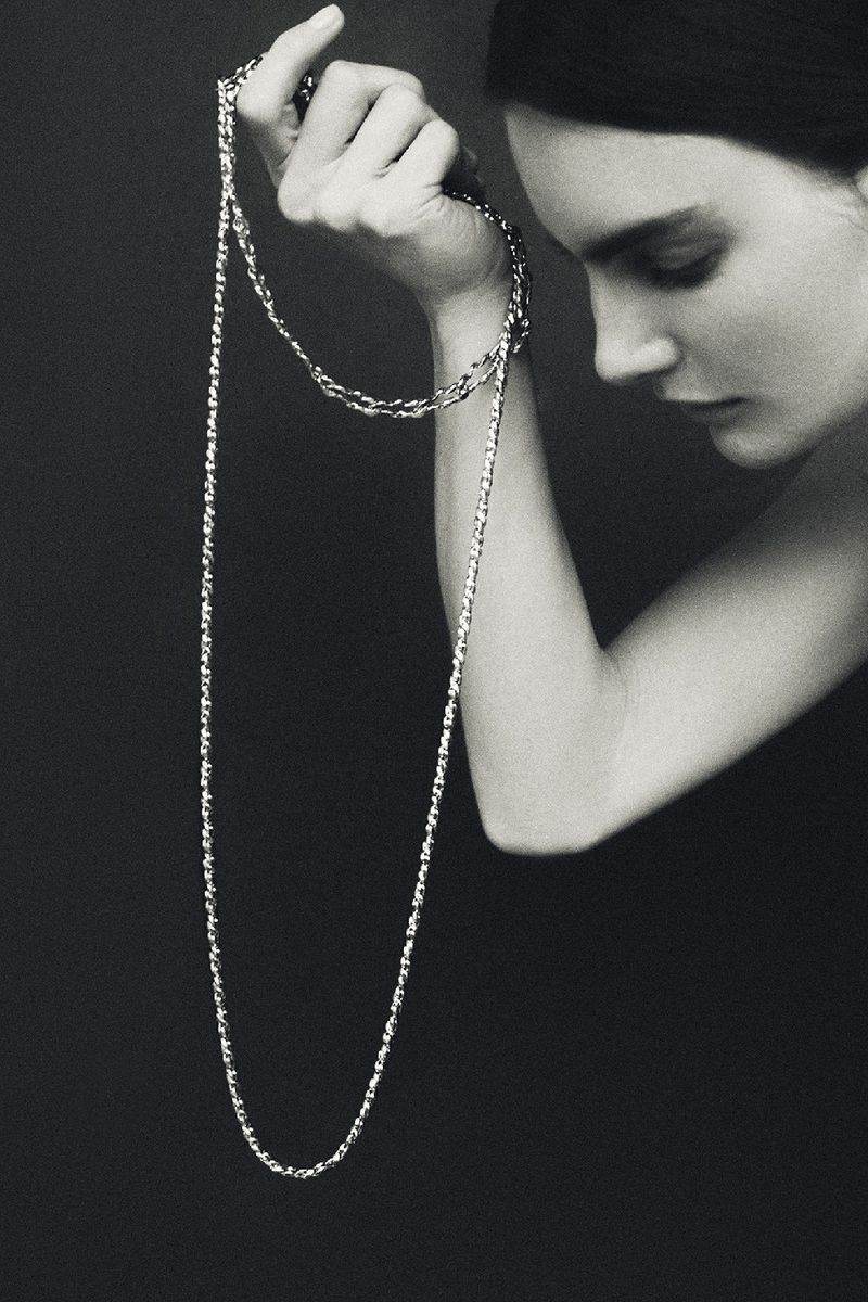 Black and white portrait of a woman holding a long metallic chain necklace looped around her wrist against a dark backdrop.