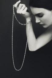 Black and white portrait of a woman holding a long metallic chain necklace looped around her wrist against a dark backdrop.