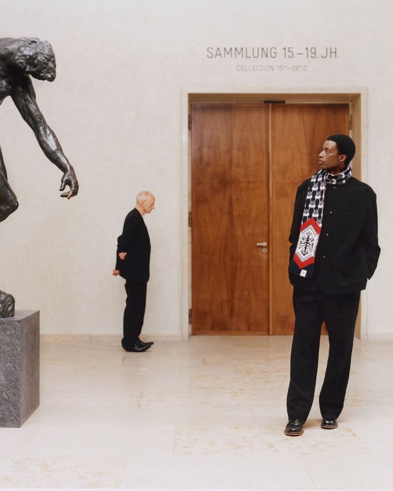 An African-American man in dark clothing stands inside a museum near a wood door, while an elderly man and a bronze statue are visible.