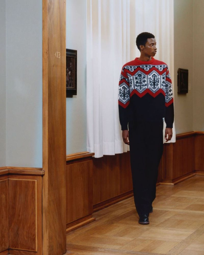 Adult male in patterned sweater and long skirt walking in an art gallery with wood floors and light-grey walls, looking to the right.
