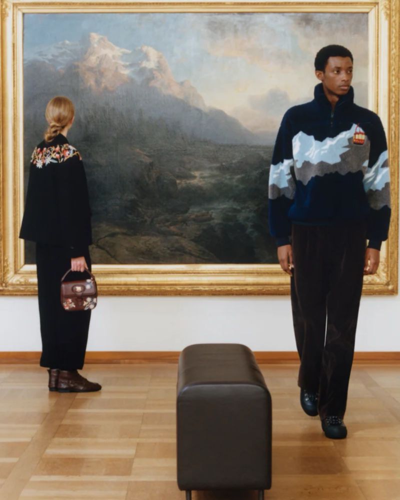 Two people in an art gallery stand before a large landscape painting while a leather bench sits in the foreground on a wood floor.