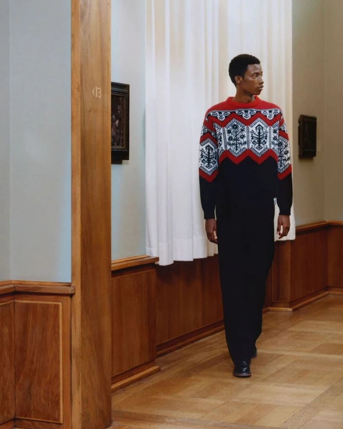 Model wearing a patterned knit sweater standing in a hallway with wooden wainscoting