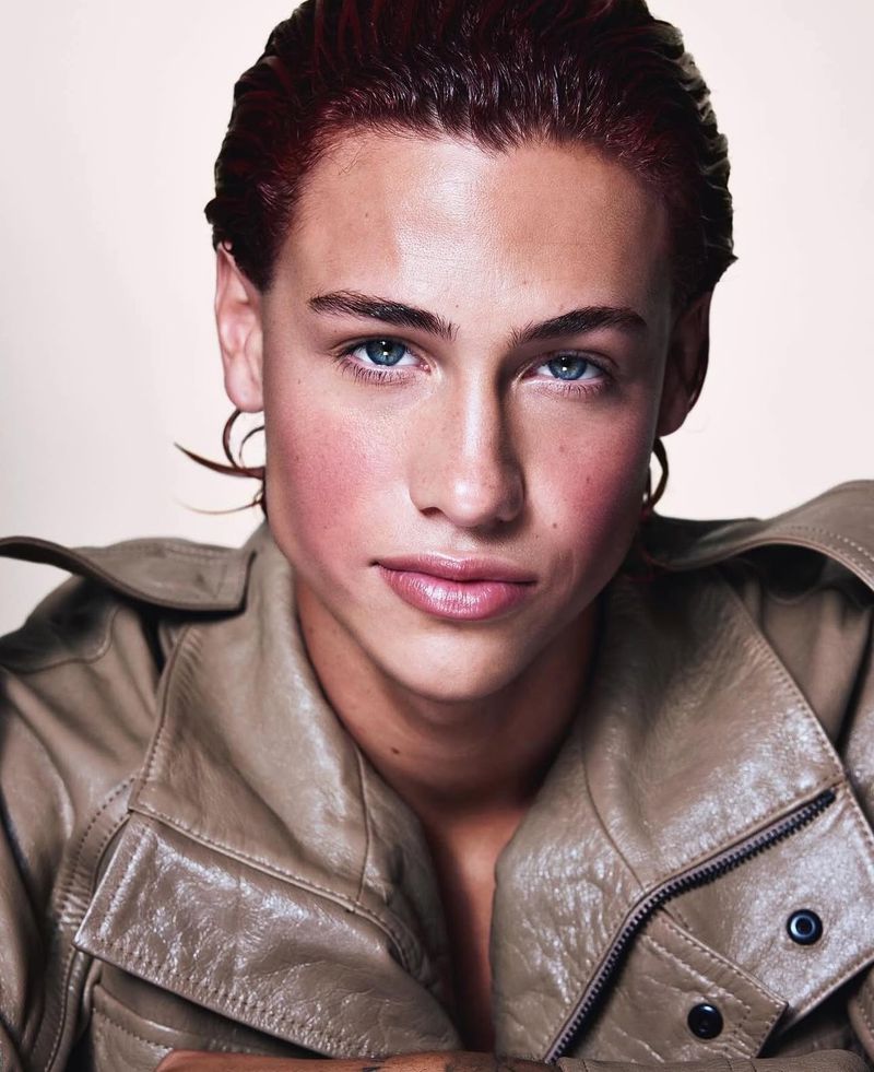 Portrait of a young adult man with light skin, light eyes, and dyed red hair. He's wearing a light brown leather jacket and posing indoors.