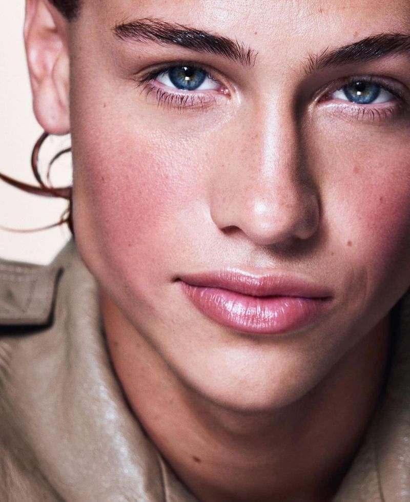 Close-up portrait of a young adult with blue eyes, tan jacket, and pink blush makeup against a neutral background in soft studio lighting.