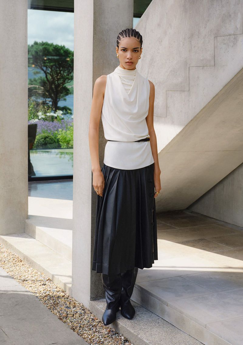 A woman poses in a white top, black skirt, and boots, standing in front of a modern house and garden, showcasing fashion and style.