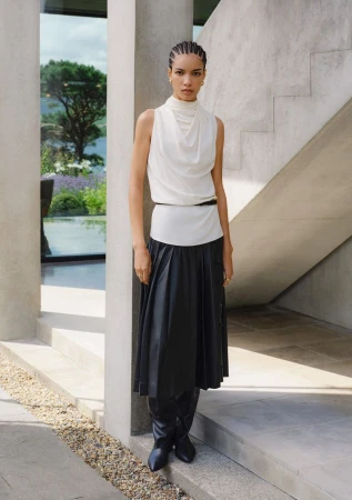 A woman poses in a white top, black skirt, and boots, standing in front of a modern house and garden, showcasing fashion and style.