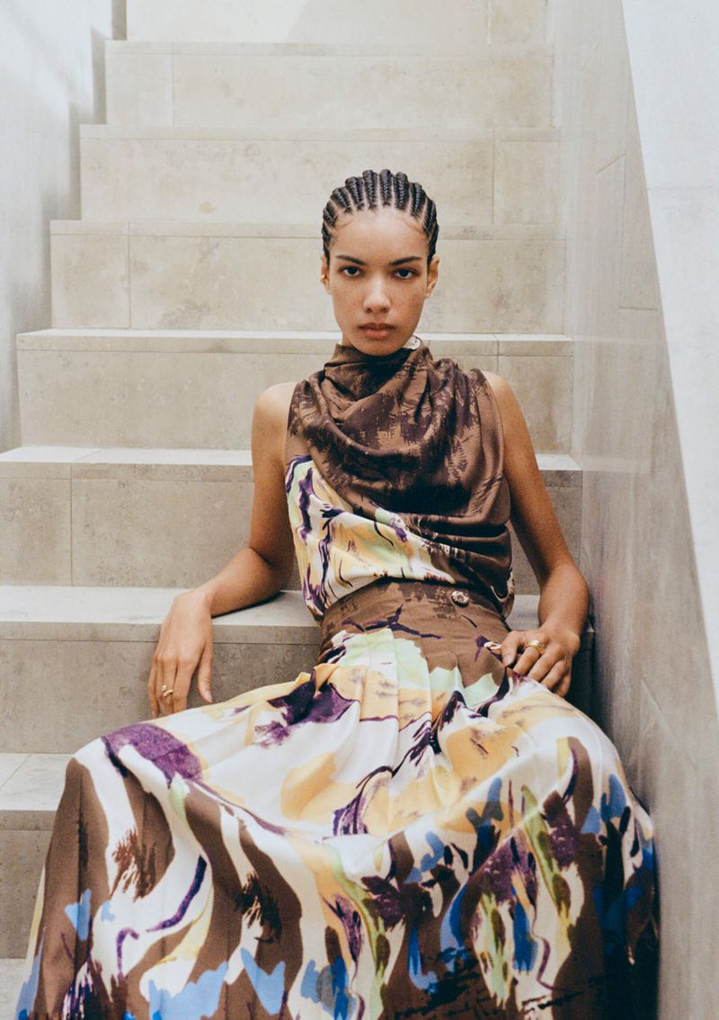 Elegant woman in a sleeveless floral dress poses on tiled steps, showcasing a chic fashion style in an indoor setting with neutral colors.