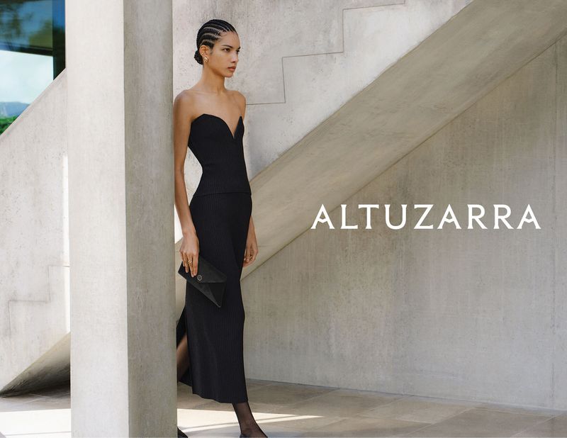 Elegant woman in black strapless dress holds clutch, posing in a contemporary building. Fashion editorial shot with brand name displayed.