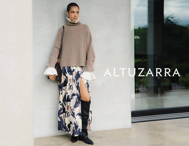 A woman in an Altuzarra sweater and a printed skirt poses in front of a neutral wall and window, holding a shoulder bag.
