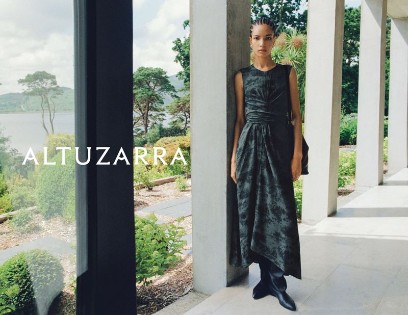 Fashion editorial featuring a solitary adult female model posing gracefully in a sleeveless midi dress amidst an outdoor setting with a colonnade.