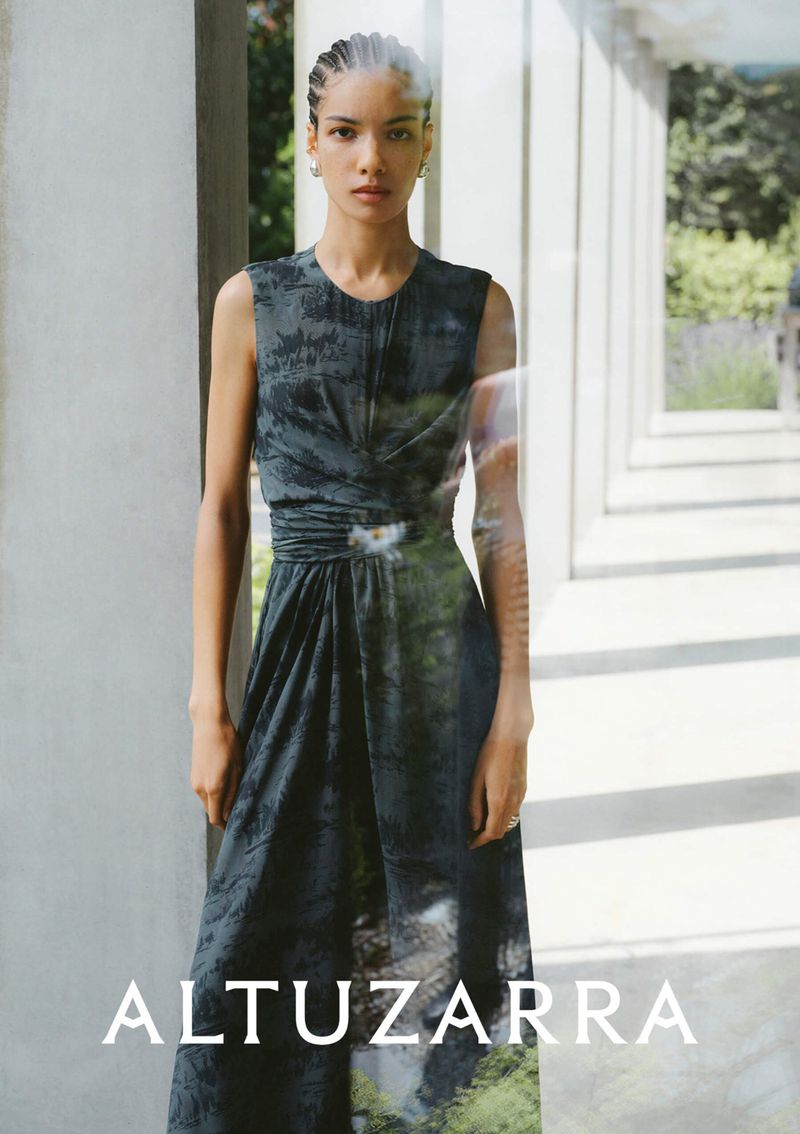 A woman in a dark dress poses in front of an outdoor hallway with white columns. The dress has a green and white print at the hem.
