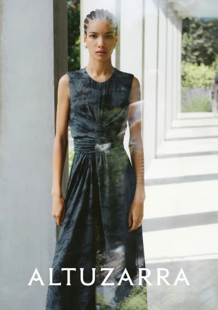 A woman in a dark dress poses in front of an outdoor hallway with white columns. The dress has a green and white print at the hem.