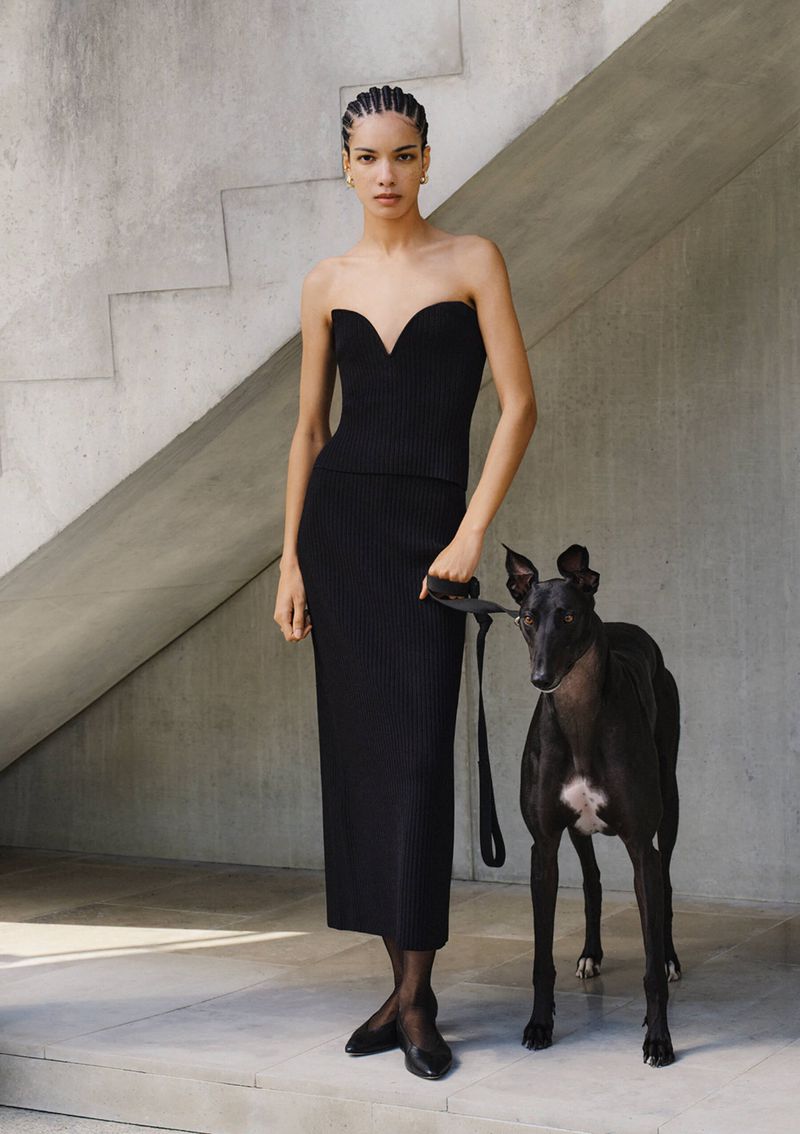 A slender adult female stands in a strapless black dress next to a sleek black dog on a leash, against a grey concrete background.