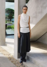 Model in a white sleeveless draped top and black pleated skirt standing on stone steps for Altuzarra