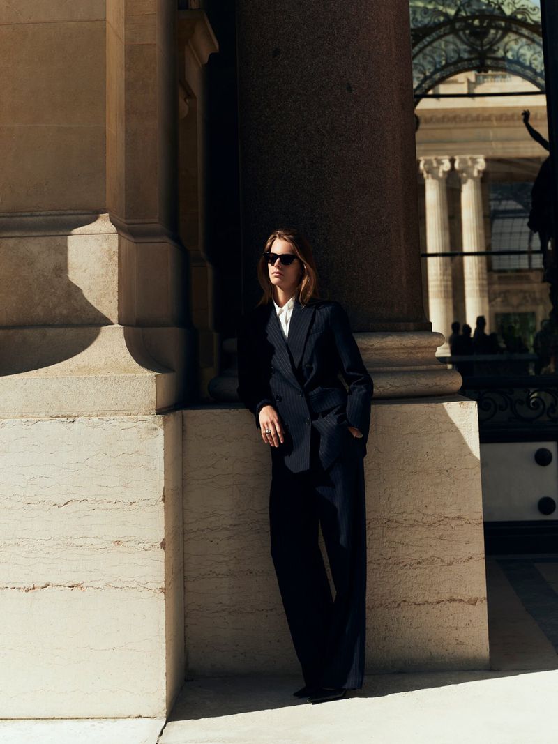 Woman in a double-breasted navy suit and sunglasses leans against a stone column in a city with classical architecture, bathed in sunlight.