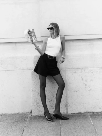 Black and white full shot of an adult woman in sunglasses, mini-skirt, top, and heels reading a magazine in an urban setting.