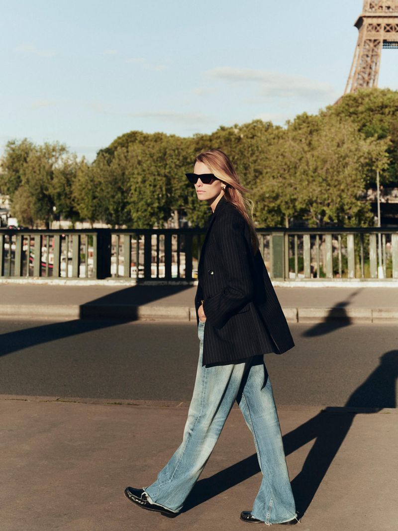 Woman walking in Paris wearing a blazer and jeans with the Eiffel Tower and trees in the background on a sunny day.