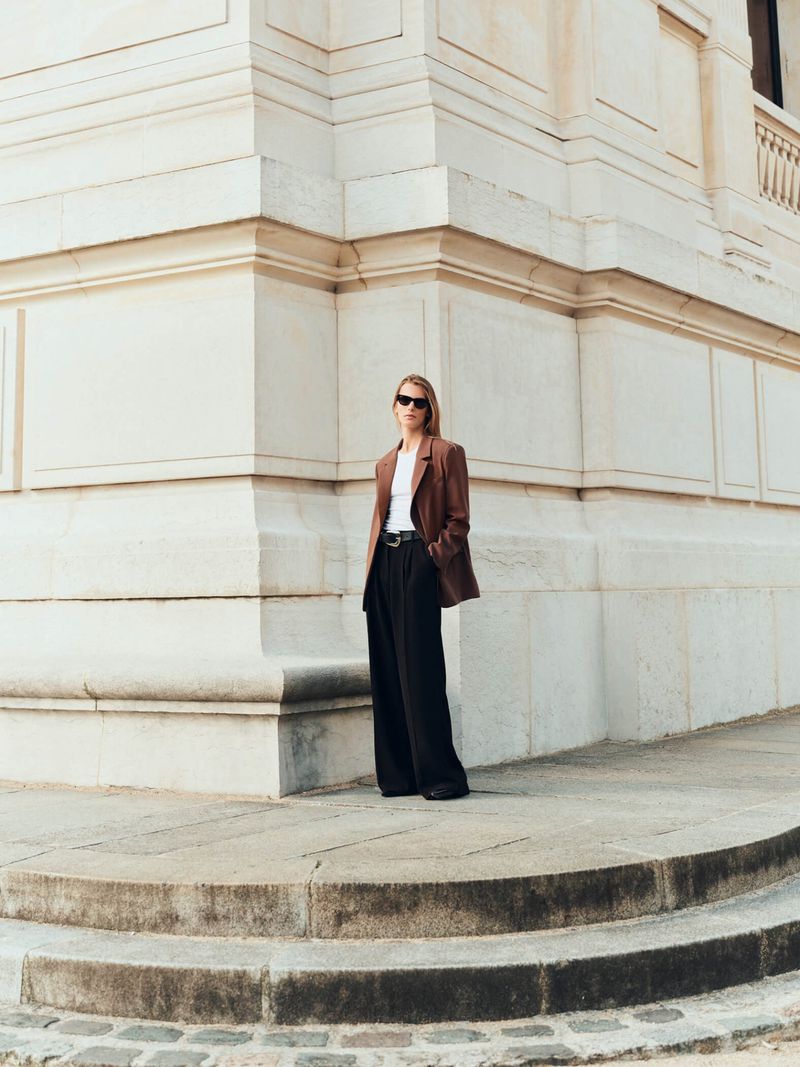 Fashionably dressed woman in brown blazer and wide-leg black pants stands near stone architecture in a full shot, wearing sunglasses.