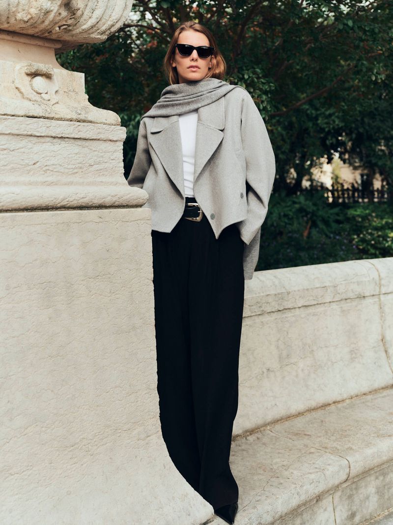 A stylish woman in sunglasses poses in front of a stone bench, wearing a gray jacket, scarf, white top, black pants, and a black belt.