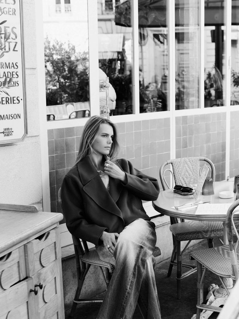 A woman in a blazer and jeans sits at a table outside a cafe, bathed in soft daylight. Reflections visible in the window.