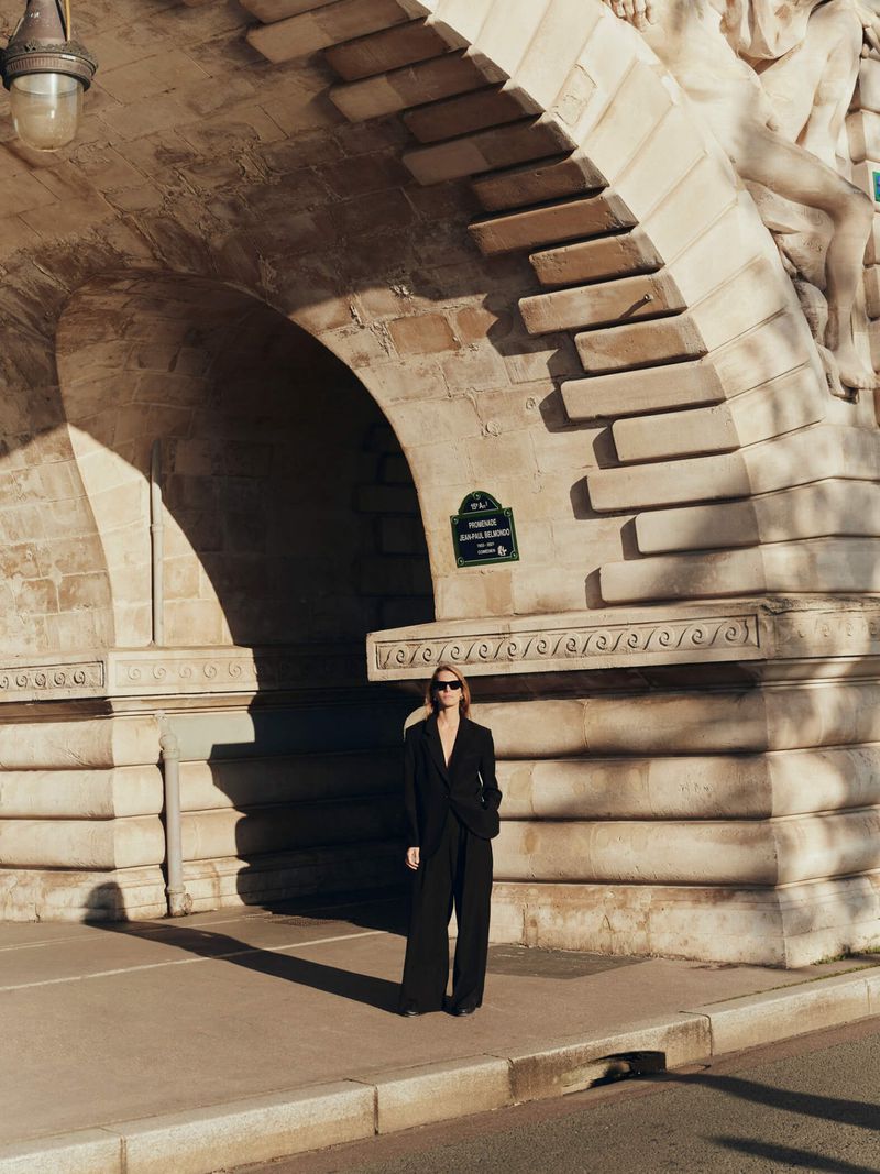 Woman in a black suit stands beneath the Pont de Bir-Hakeim bridge in Paris, on a sunny day, showcasing architecture and shadows.