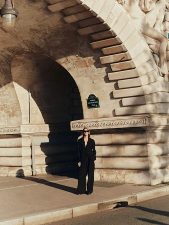 Woman in a black suit stands beneath the Pont de Bir-Hakeim bridge in Paris, on a sunny day, showcasing architecture and shadows.