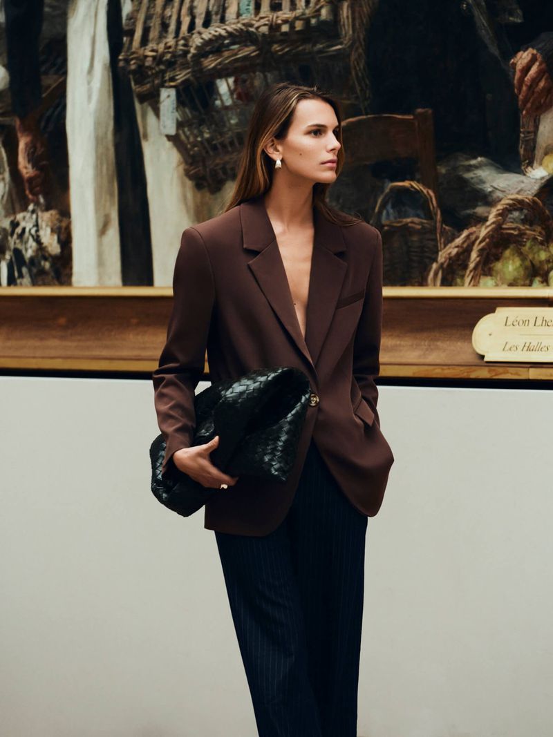 A woman in a brown blazer poses in front of a painting, holding a black woven purse, wearing a navy skirt, and looking off to the side.