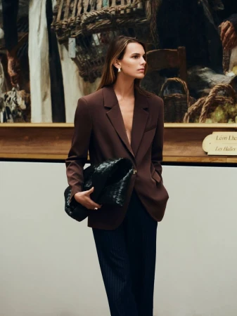 A woman in a brown blazer poses in front of a painting, holding a black woven purse, wearing a navy skirt, and looking off to the side.
