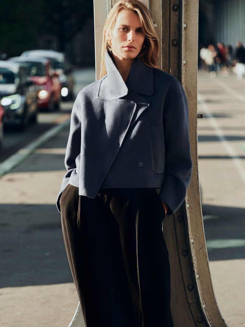A woman stands on a bridge in stylish dark pants and a blue jacket, cars visible in the background.