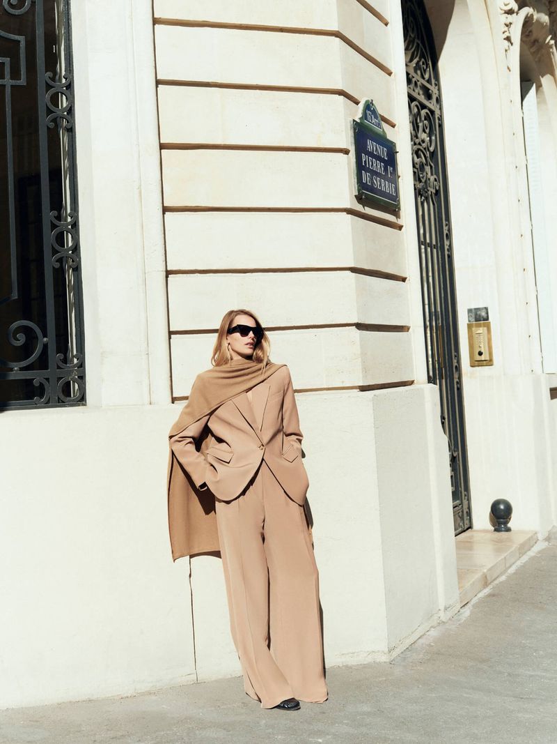 Adult woman in a tan pantsuit and sunglasses poses on a city street with a light building facade backdrop on a sunny day.