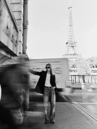 Model in sunglasses wearing a dark jacket and jeans standing outdoors in Paris with the Eiffel Tower in the background.