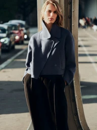 Model leaning against a structural metal pillar wearing a navy blue wool jacket and dark trousers on an urban street.