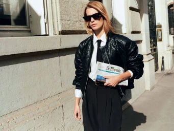 Model in dark sunglasses wearing a black quilted bomber jacket and white shirt holding a newspaper outdoors
