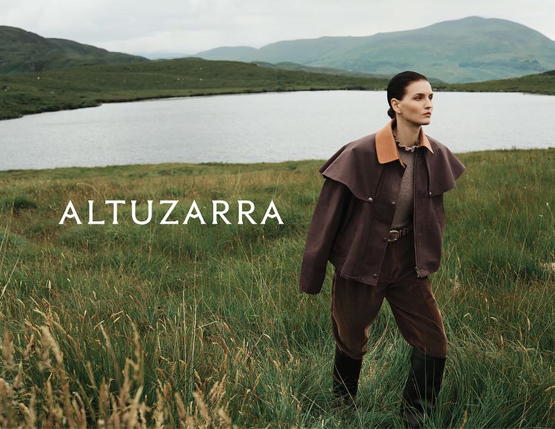 A woman in a brown jacket and pants walks through a field of tall grass, with a lake and mountains in the background, fashion editorial.