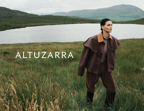 A woman in a brown jacket and pants walks through a field of tall grass, with a lake and mountains in the background, fashion editorial.