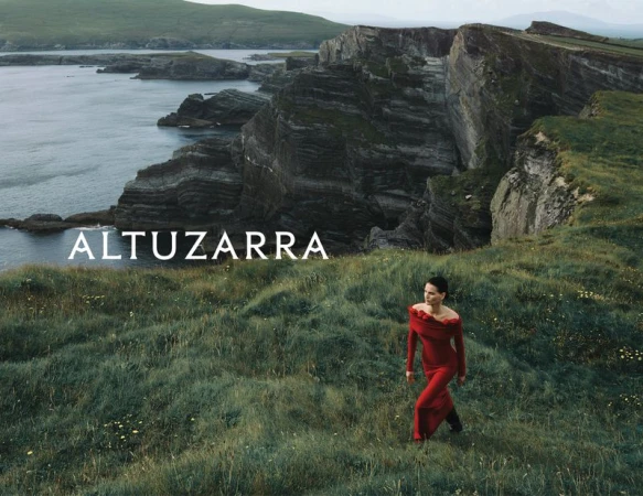 A woman in a red dress walks on a green hillside with a rocky coast in the distance under an overcast sky. Fashion editorial.
