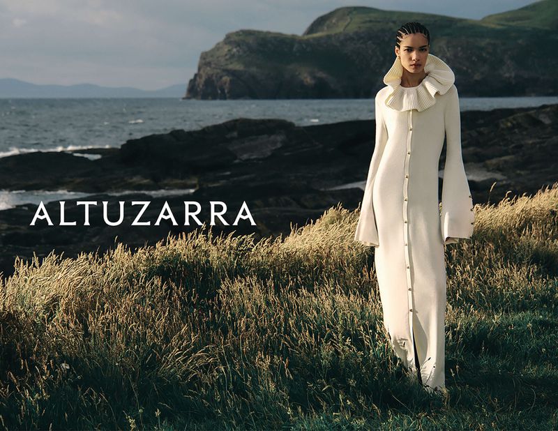 A woman stands in a long, white dress with a ruffled collar, posing in an outdoor setting with tall grasses and a coastal backdrop.