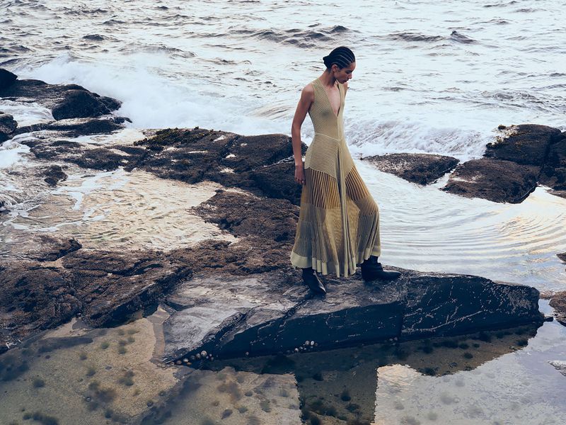 Adult female in a flowing dress stands on ocean rocks, waves behind. Moody outdoor fashion shot with natural light and shallow depth of field.