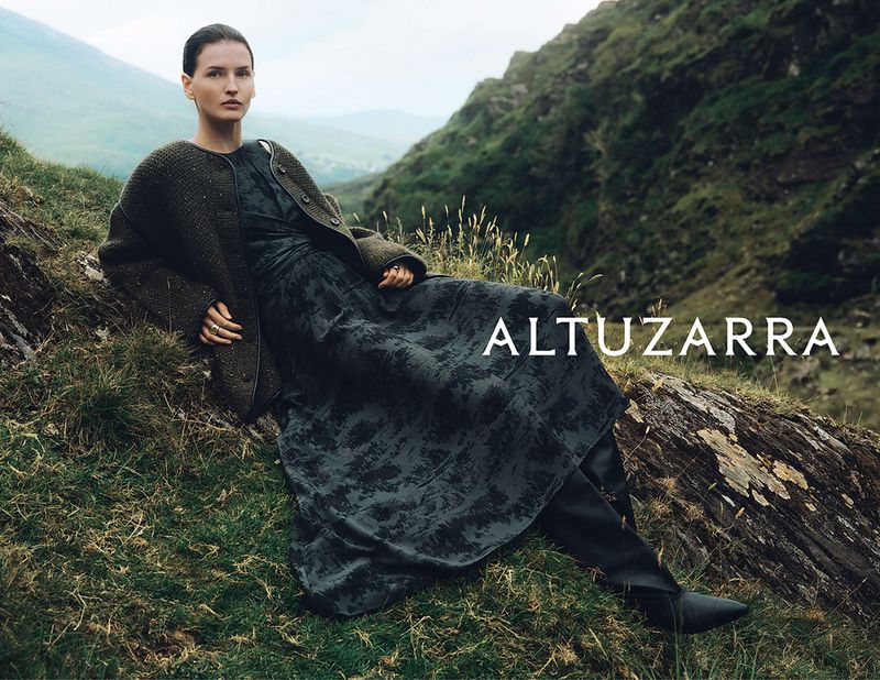 A woman in a dress and jacket is posed outdoors with mountainous scenery, creating a fashion editorial look with natural and stylish elements.